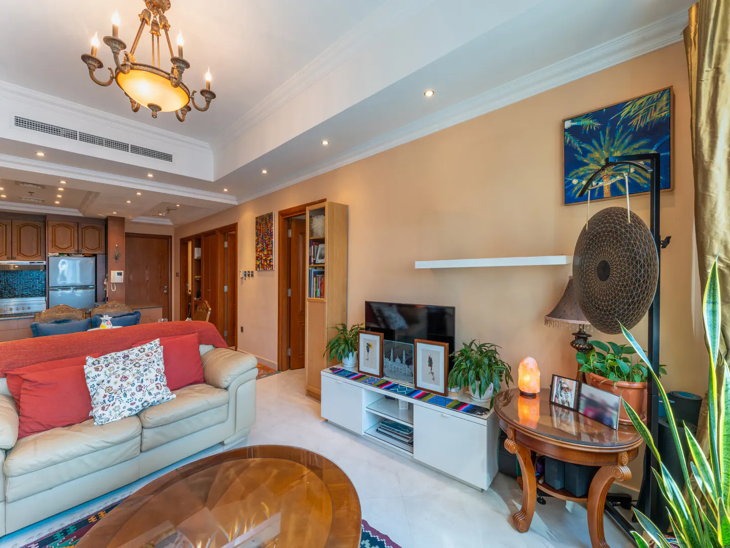 A living room with a beige leather sofa, red pillows, a glass coffee table, and a white TV stand with plants. A gong and a palm tree painting are on the wall.