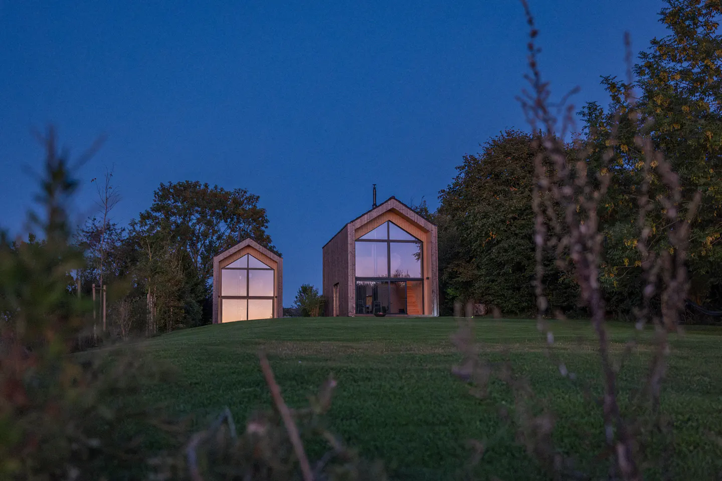 Two modern, wood-clad homes with large windows sit on a green lawn at dusk, surrounded by trees.