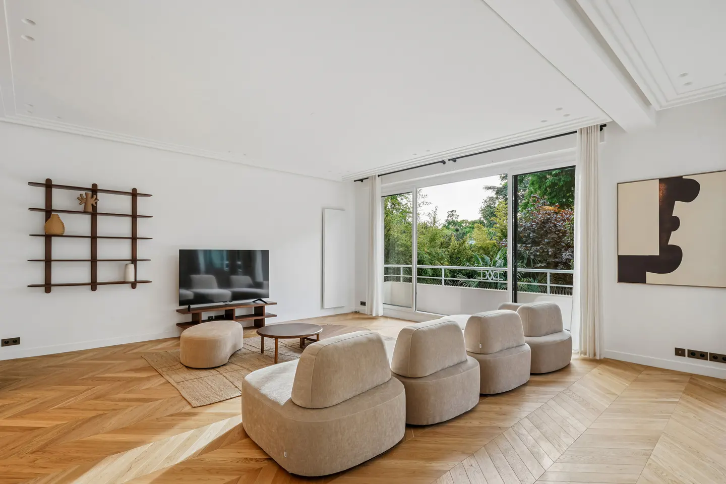 Bright living room with herringbone wood floors, beige modular seating, TV, and balcony overlooking green trees.