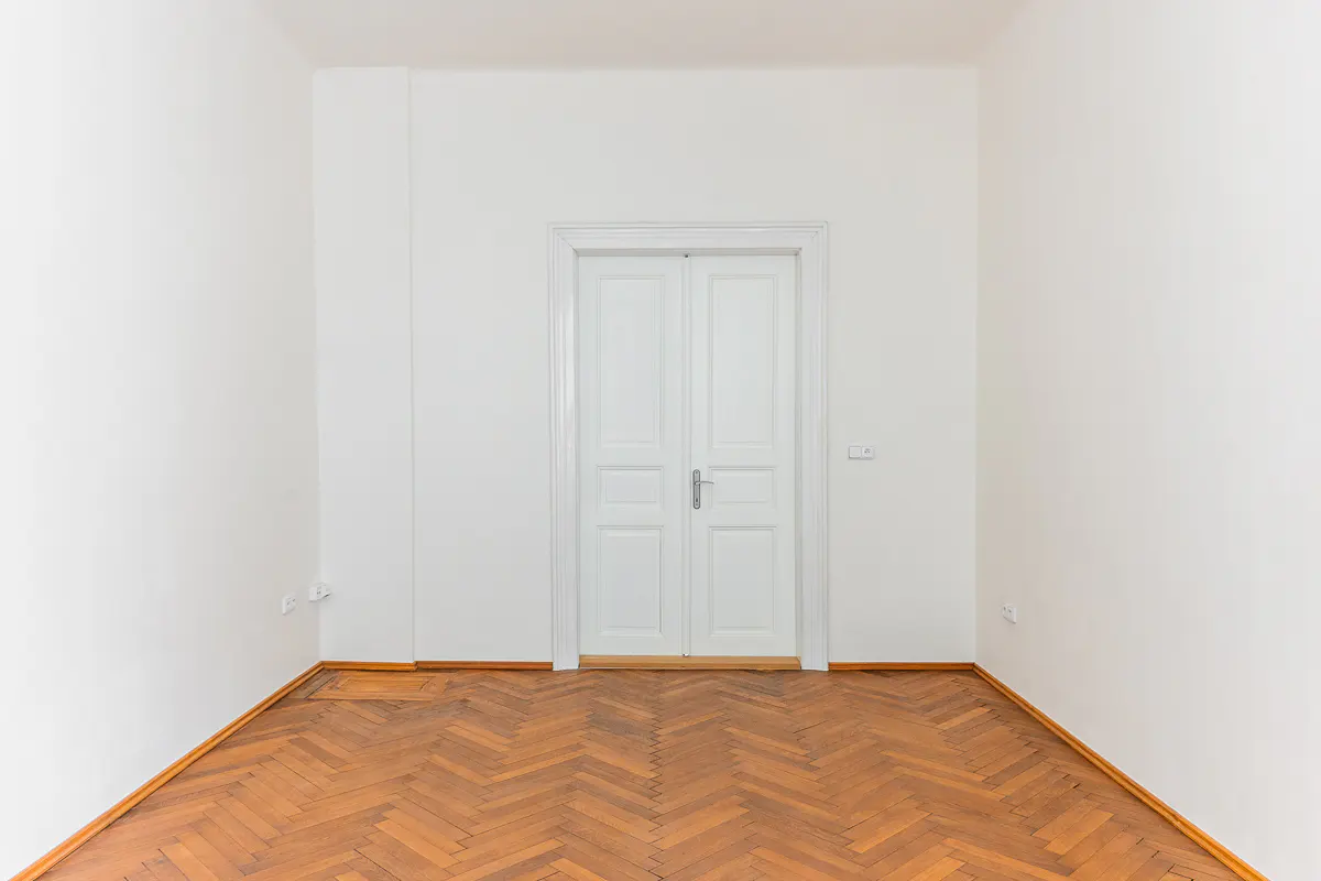 Empty room with white walls, herringbone wood floor, and a centered white double door.