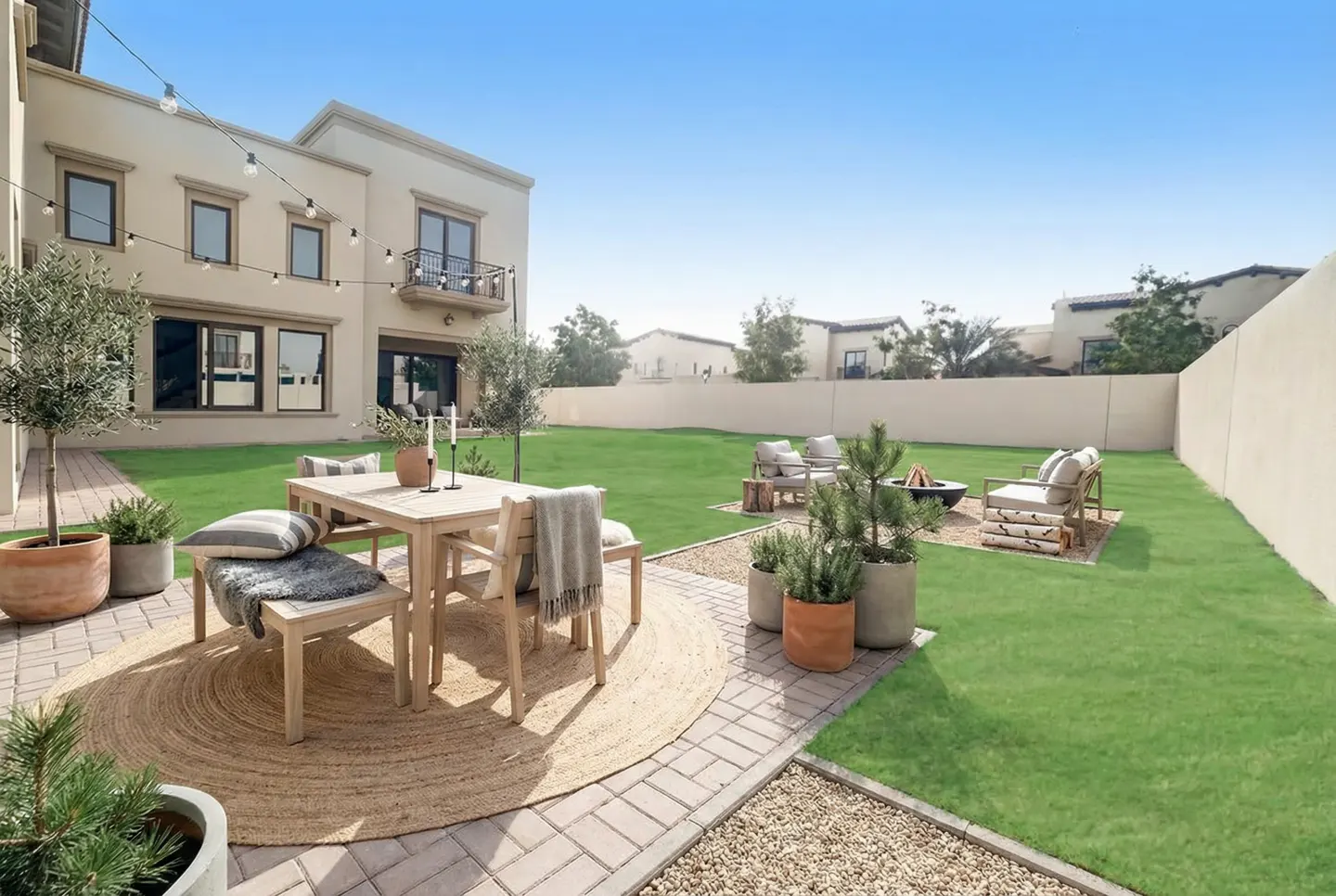 Backyard patio with a wooden table, chairs, and a round rug on a brick patio. A fire pit and seating area are on the green lawn.