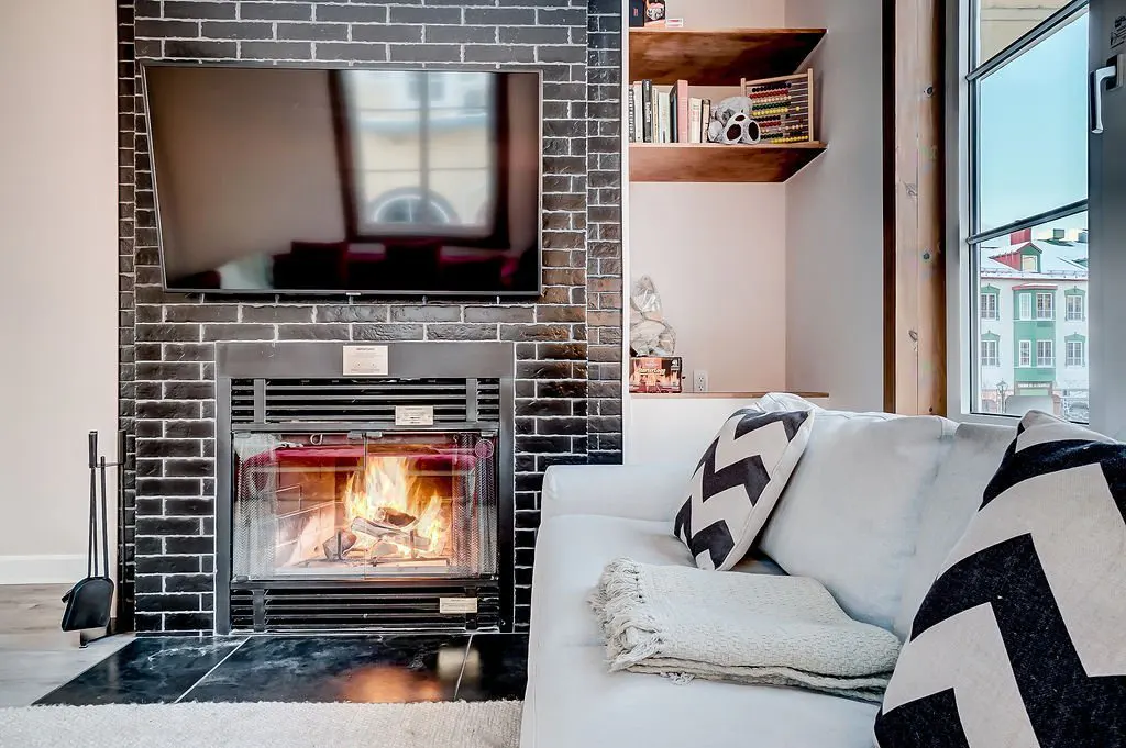 Living room with a black brick fireplace, TV, and white sofa with chevron pillows. A window shows a building outside.