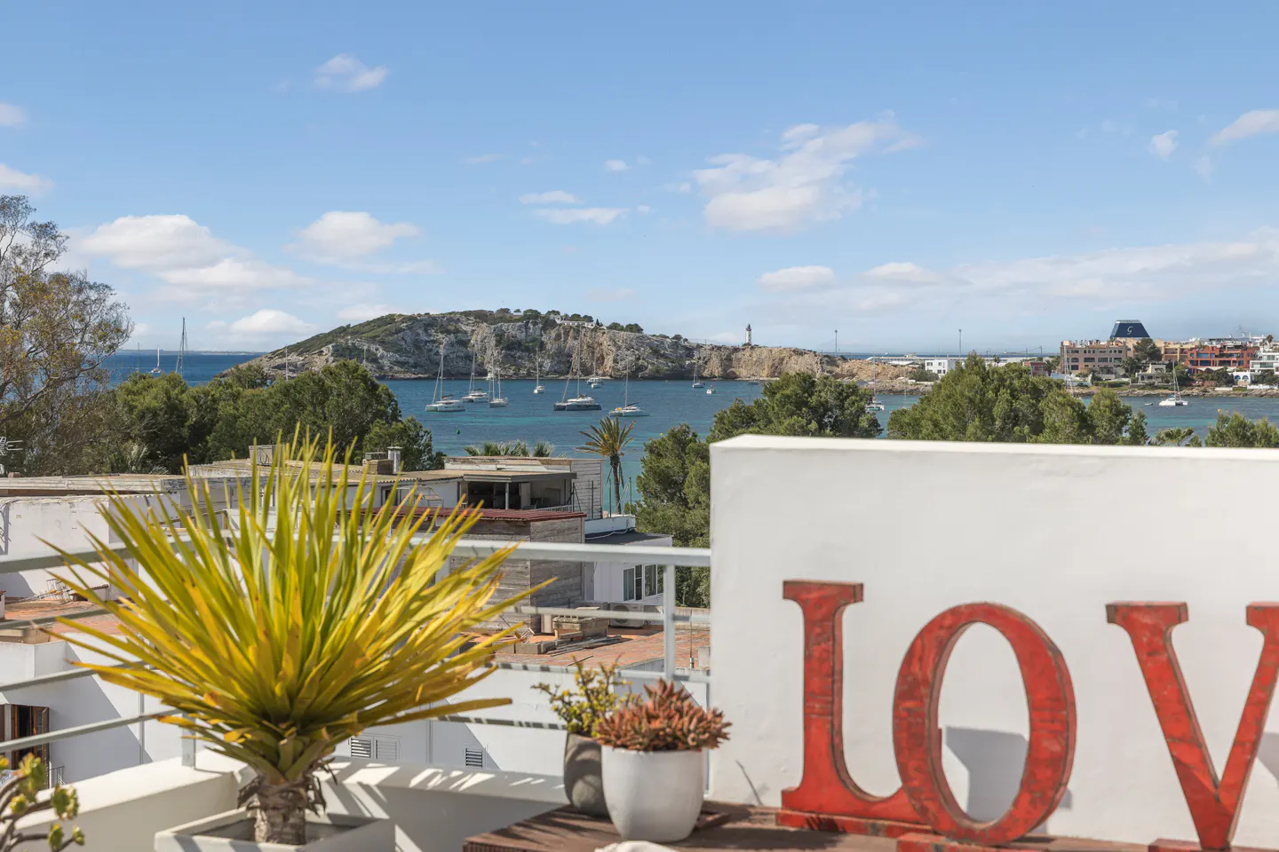 Balcony view of a blue sea with sailboats, green trees, and a distant island under a blue sky. A "LOVE" sign and potted plants are in the foreground.