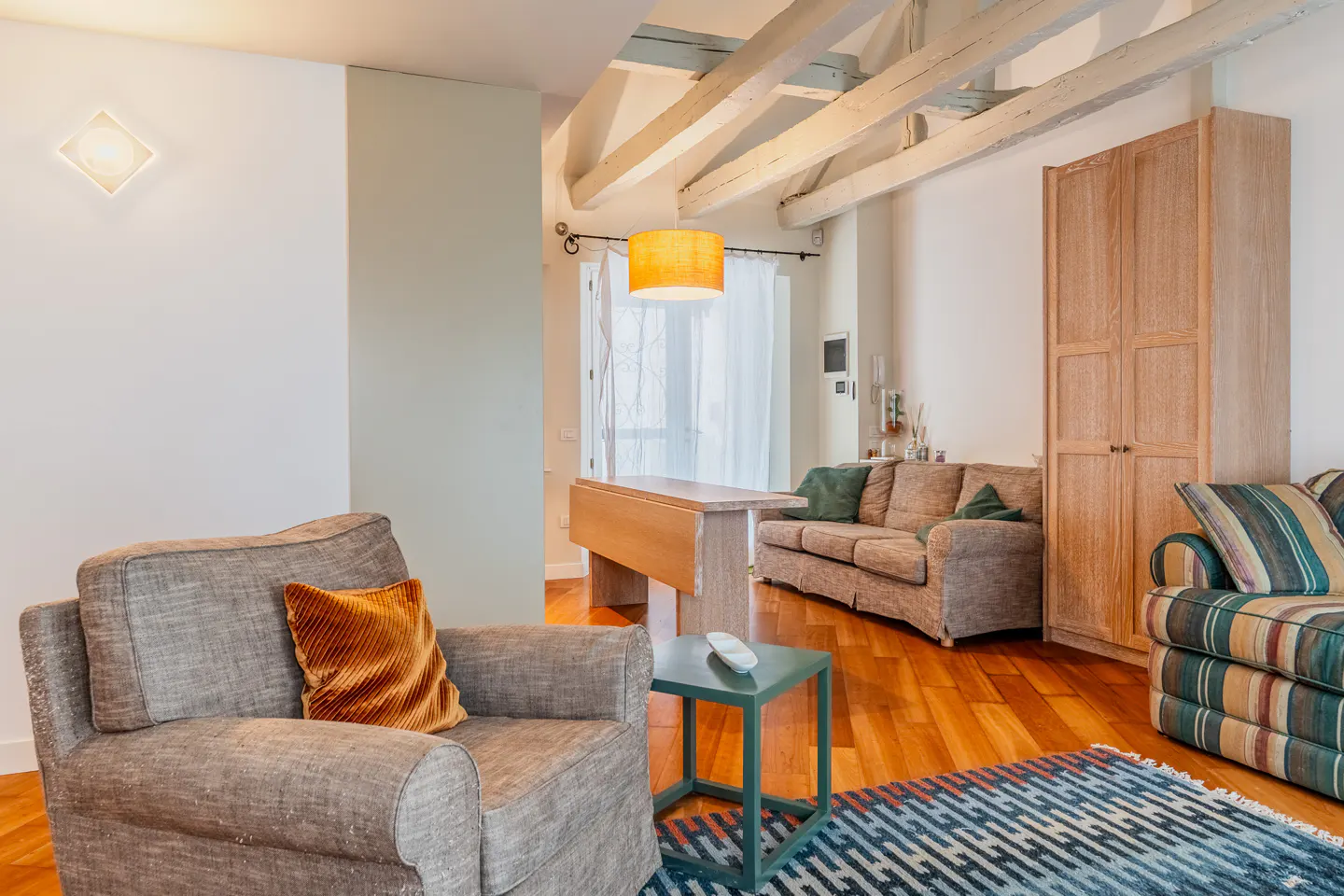 Living room with grey armchair, orange pillow, wooden beams, sofa, tall cabinet, and blue patterned rug on hardwood floors.