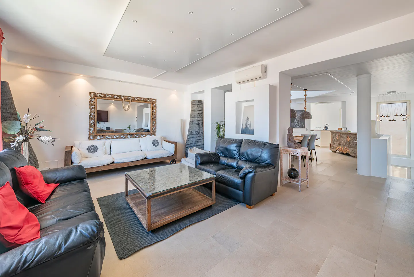 Bright living room with black leather sofas, red pillows, and a wooden coffee table on a gray rug. A large mirror hangs above a white sofa.