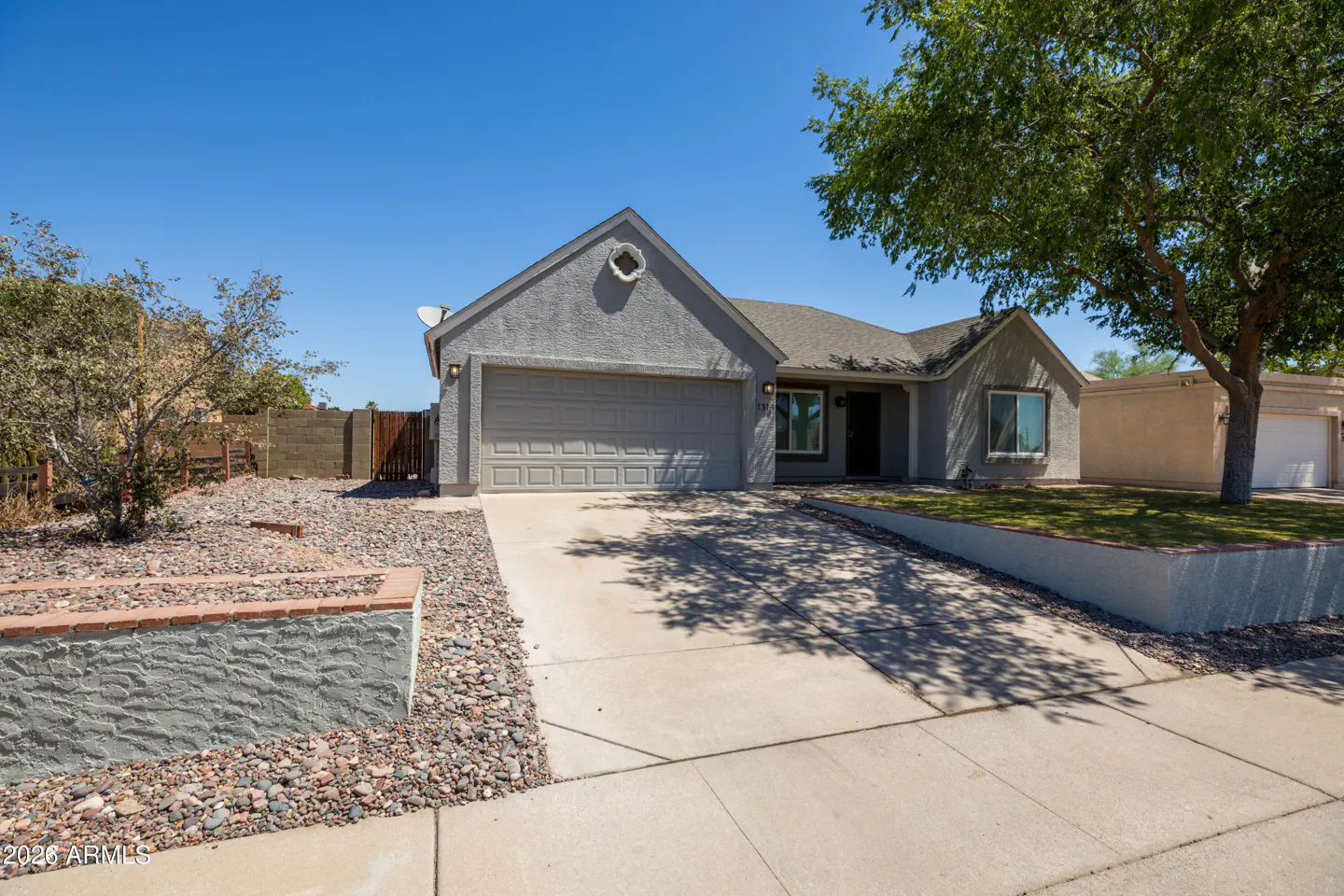 Single-story gray stucco home with a two-car garage, concrete driveway, and desert landscaping under a clear blue sky.