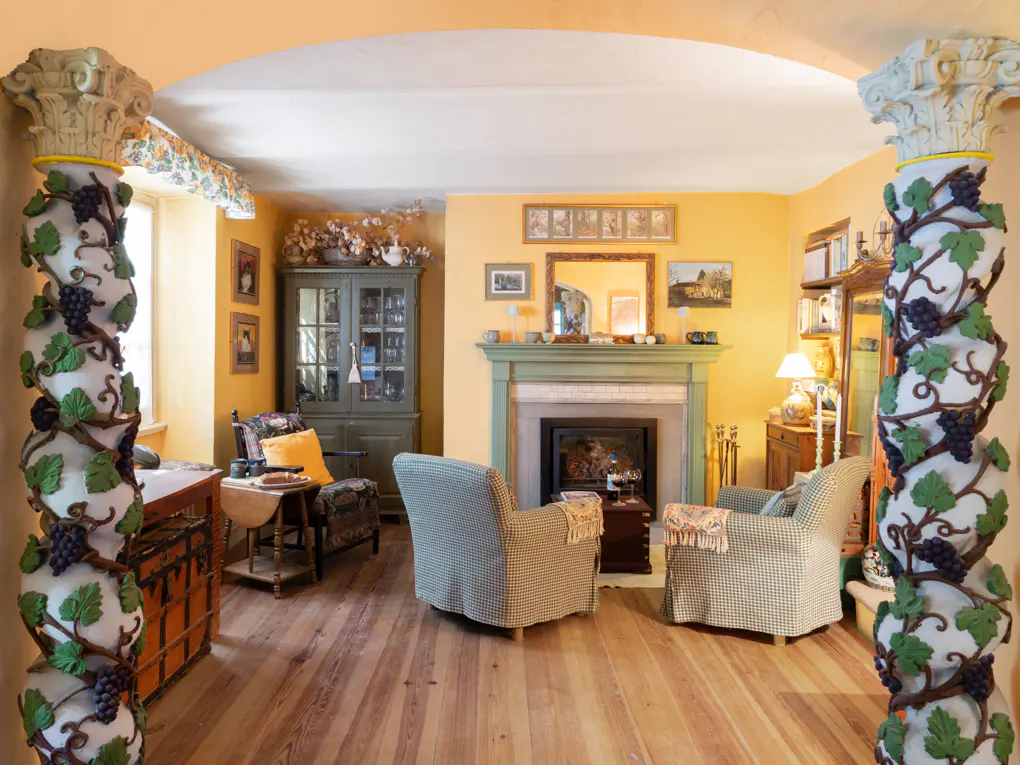 Living room with yellow walls, wood floors, and two armchairs facing a fireplace. Columns with grape vine decor frame the view.