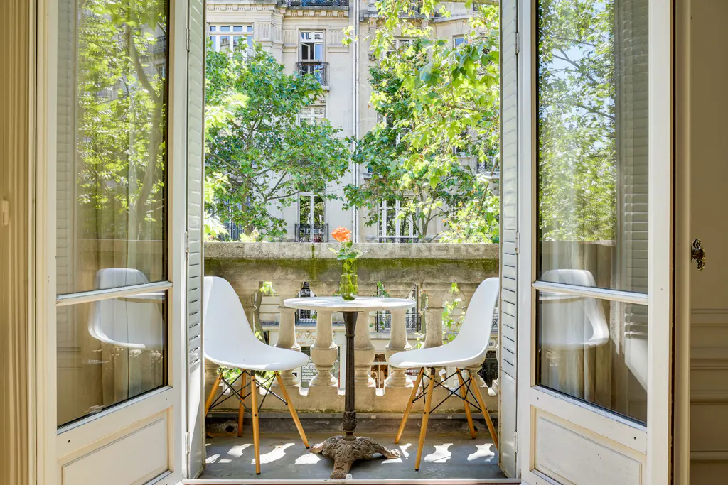 Open French doors lead to a balcony with two white chairs and a small table with a rose in a vase.