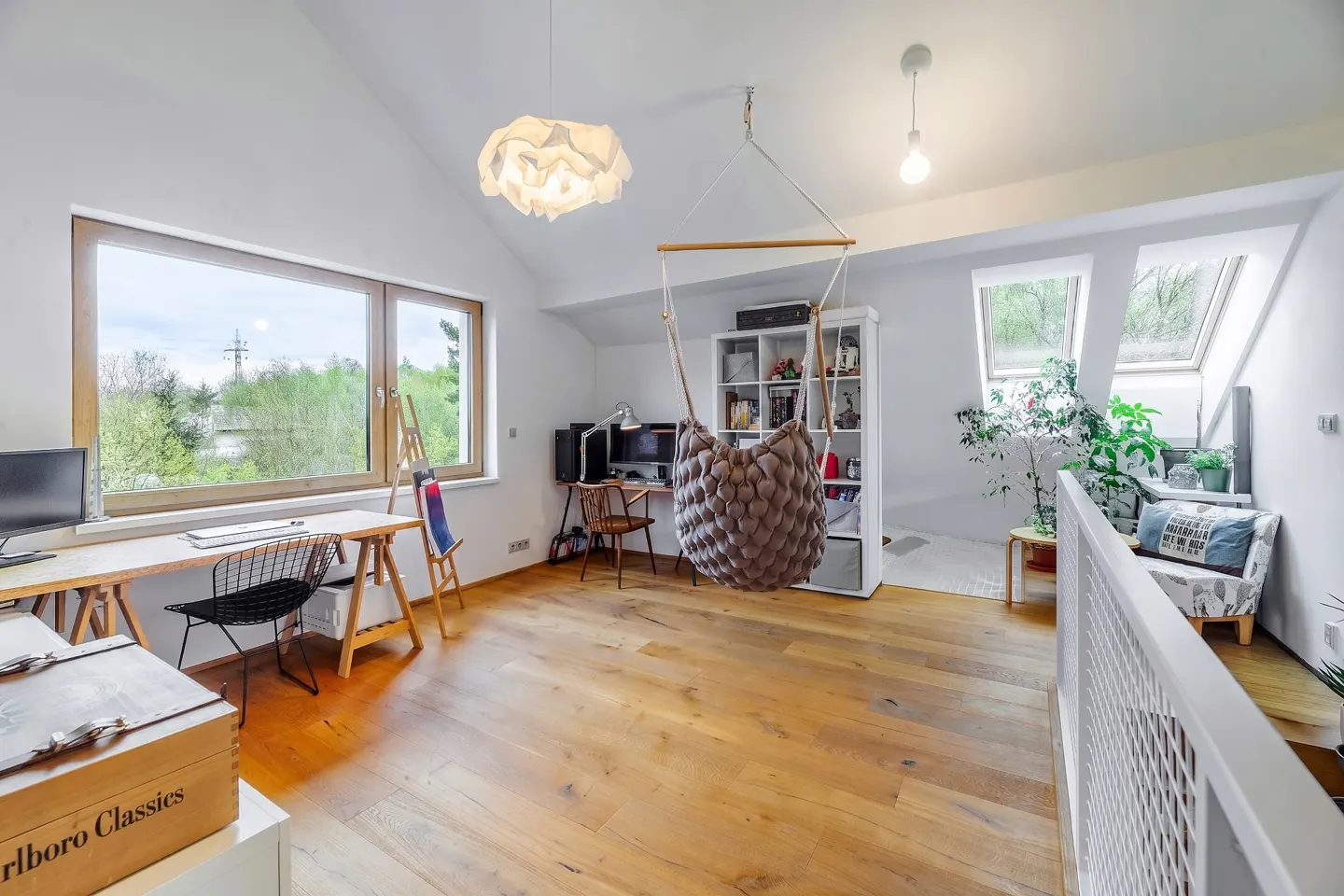 Bright attic room with wood floors, white walls, and a large window. A hanging chair, desk, and bookcase furnish the space.
