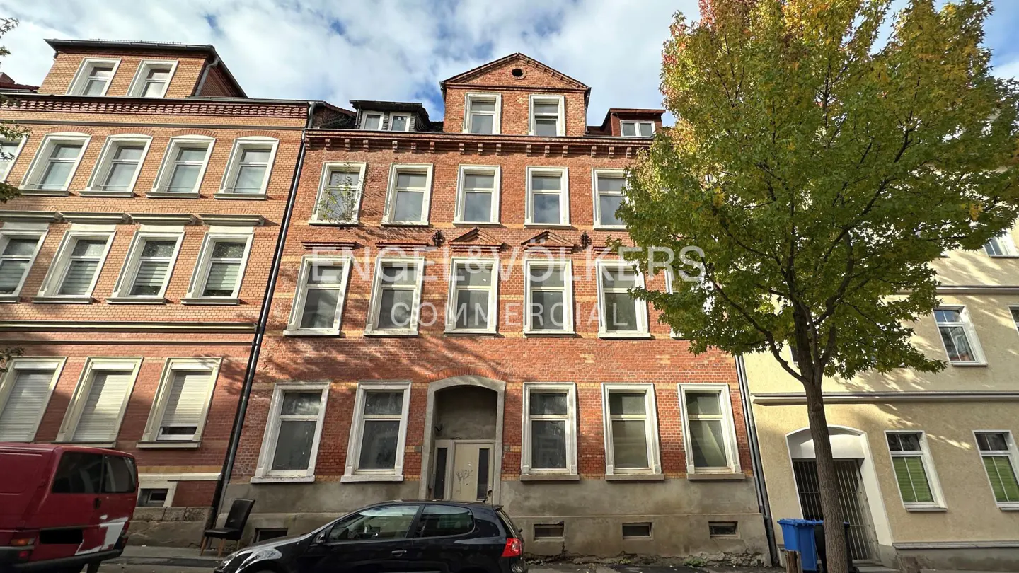 A three-story red brick building with white-framed windows and a tree on the right. A black car is parked in front.