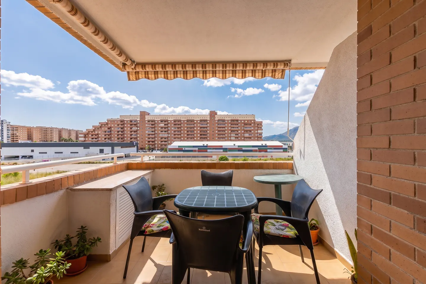 Balcony with black table and chairs, brick wall, and city view under a blue sky.