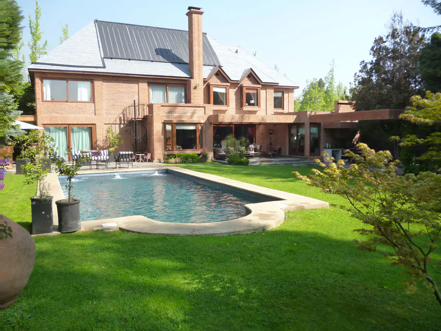 A brick house with a gray roof overlooks a pool and green lawn. Trees surround the property.