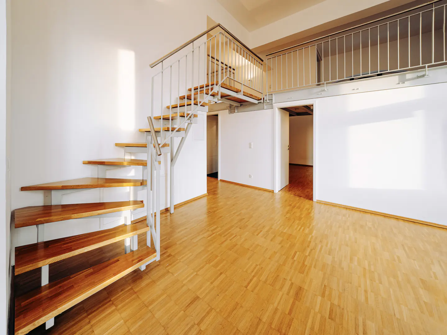Bright, modern apartment interior with wood floors, white walls, and a staircase leading to a loft area with metal railings.