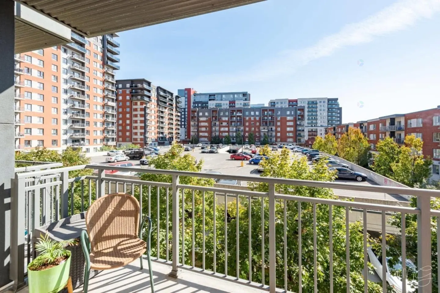 View from a balcony with a chair and plant, overlooking a parking lot and apartment buildings on a sunny day.