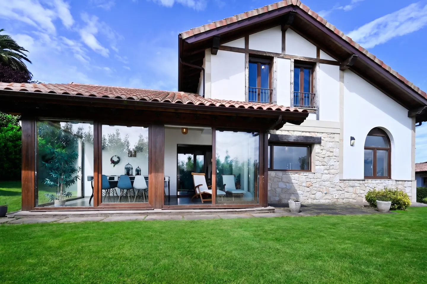 Exterior view of a two-story house with a green lawn, stone accents, and a sunroom with glass walls.