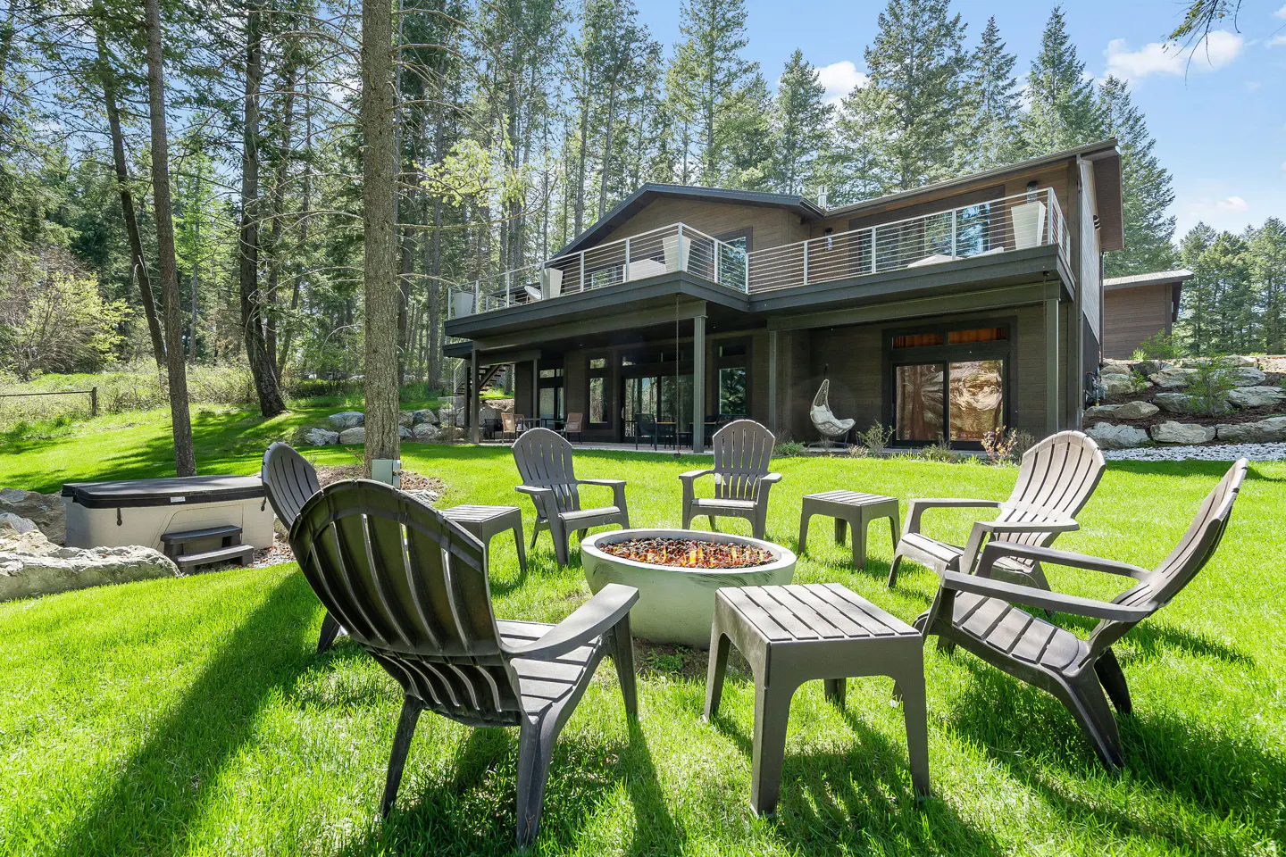 Backyard of a modern brown house with a fire pit surrounded by chairs and small tables on a green lawn. Tall trees in the background.