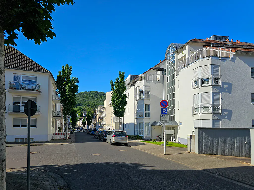 A sunny street scene with white apartment buildings, parked cars, and a green hill in the distance under a clear blue sky.