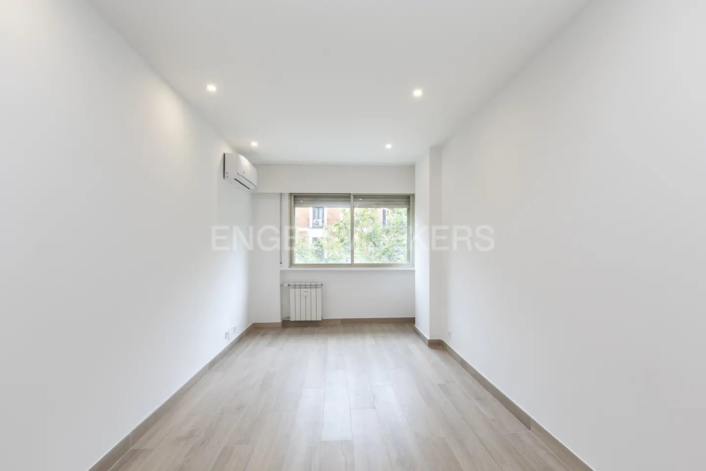 Bright, empty room with light wood floors, white walls, and a window showing trees. A radiator and AC unit are visible.