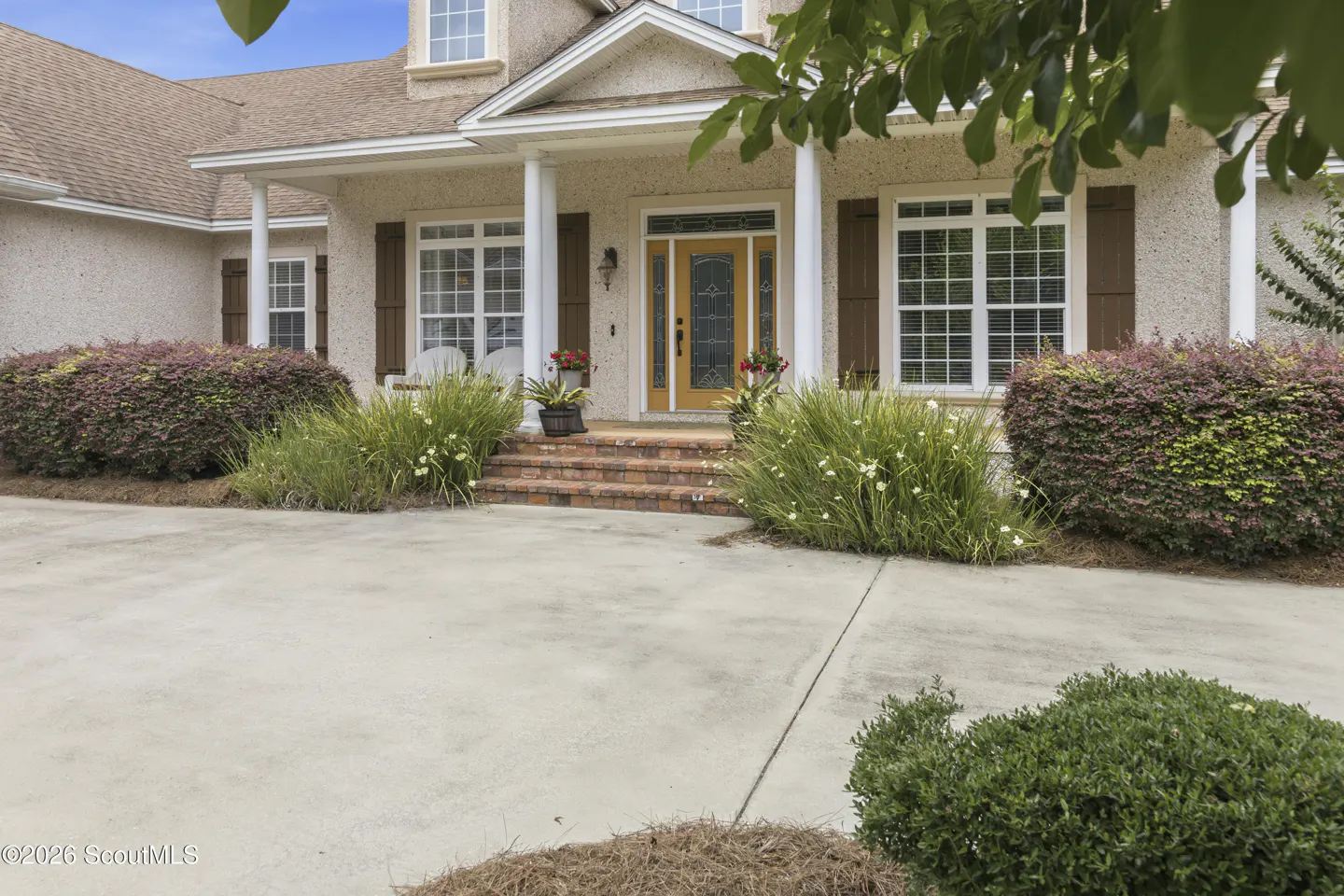 Beige stucco house with brown shutters, white trim, and a yellow front door. Brick steps lead to the porch. Bushes line the front.