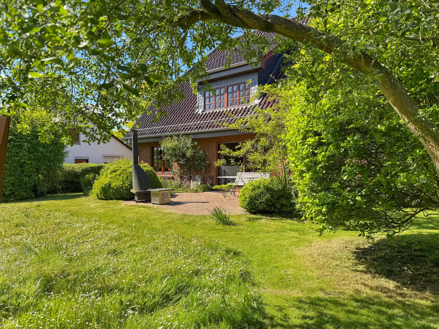 A brick house with a red tile roof is surrounded by green trees and grass. A metal chimney stands on a stone patio.