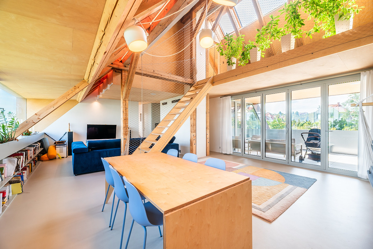 Bright, modern living room with wood beams, a long table with blue chairs, and a blue sofa. A staircase leads to a loft with plants.