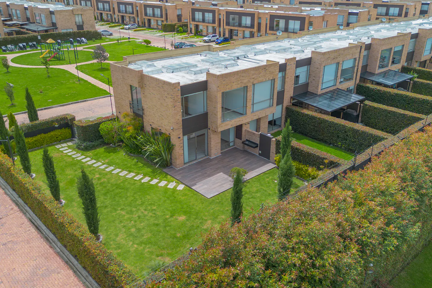 Aerial view of a modern brick townhouse with a private backyard, green lawn, stone path, and tall hedges in a neighborhood.