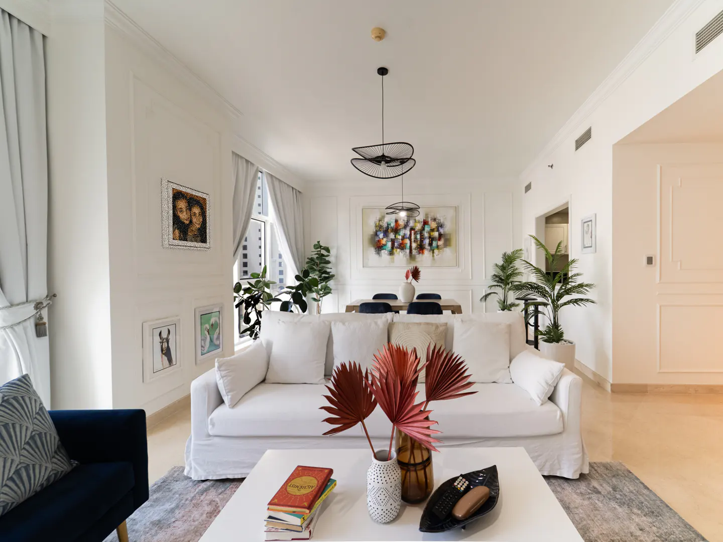 Bright living room with white sofa, coffee table with books and decor, and dining area with abstract art. Plants add a touch of nature.