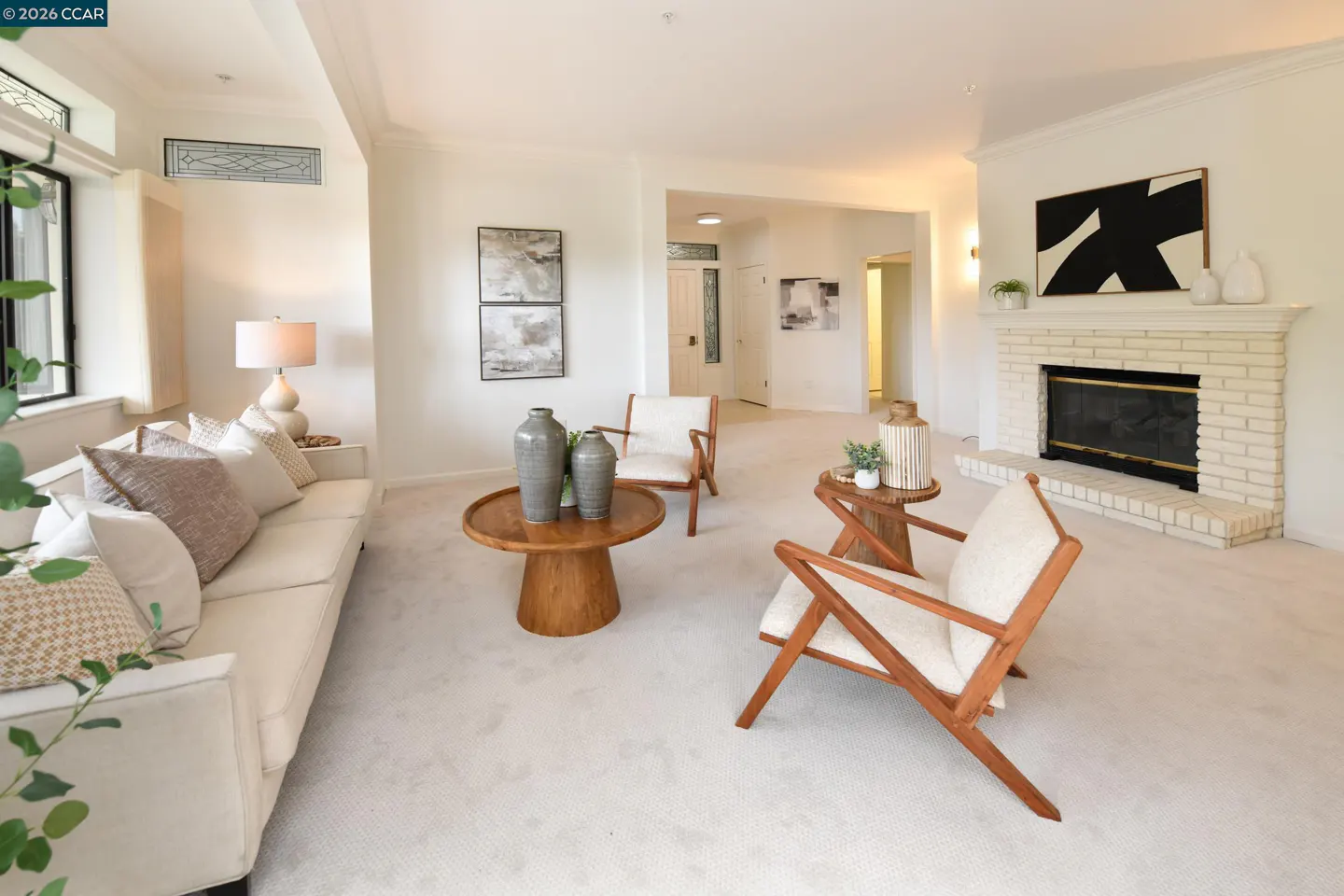 Bright living room with white walls, carpet, and furniture. A brick fireplace with black and white art is visible. Two wooden chairs and tables are in the center.