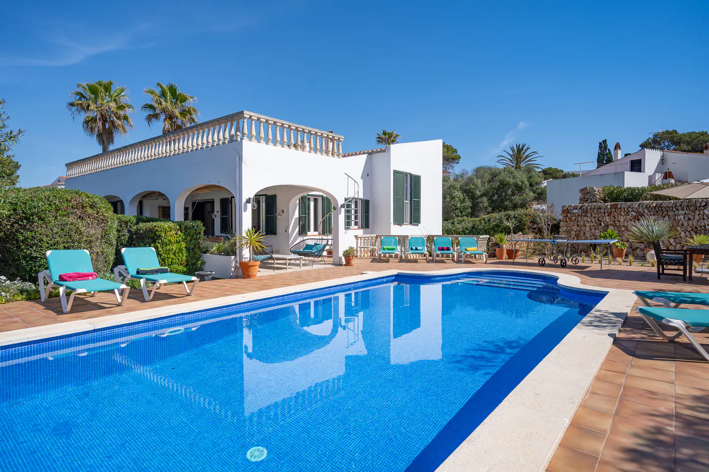 Bright sunny view of a white villa with a blue tiled pool, lounge chairs, and palm trees.