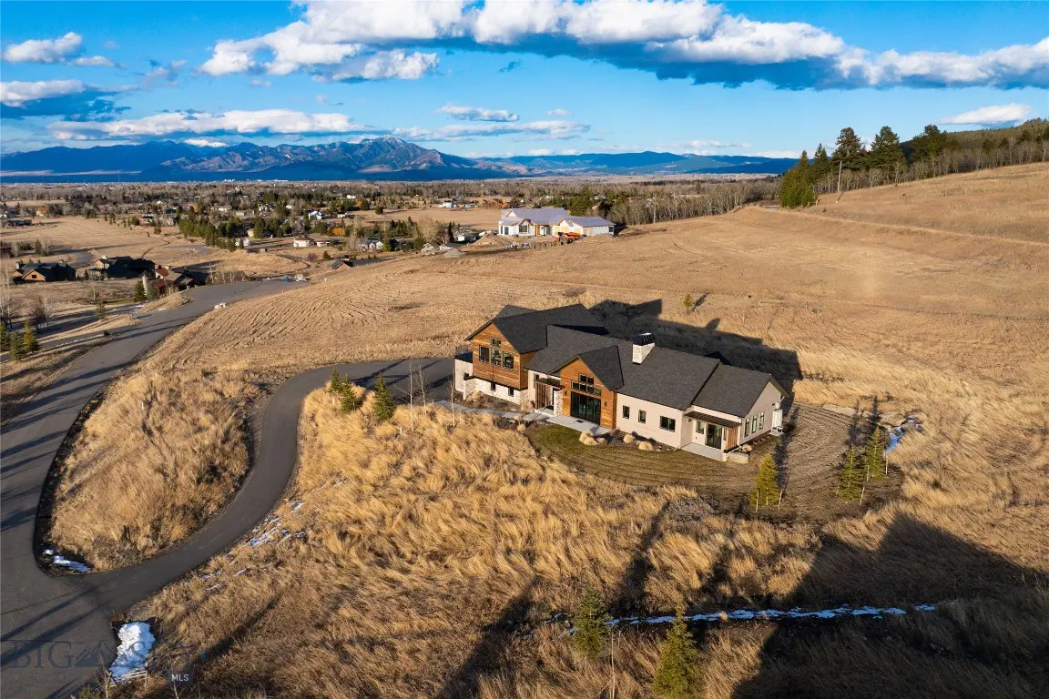 Aerial view of a modern house with a dark roof on a grassy hill, mountains in the background, and a blue sky with clouds.
