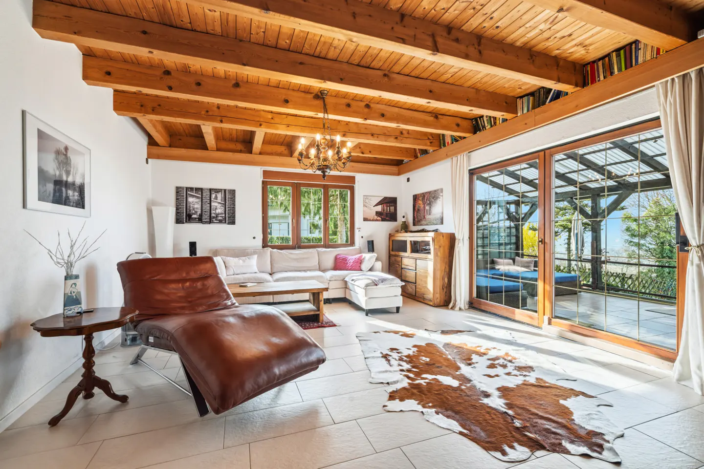 Bright living room with wood-beamed ceiling, chandelier, and large windows. A brown leather chaise lounge and cowhide rug add texture.