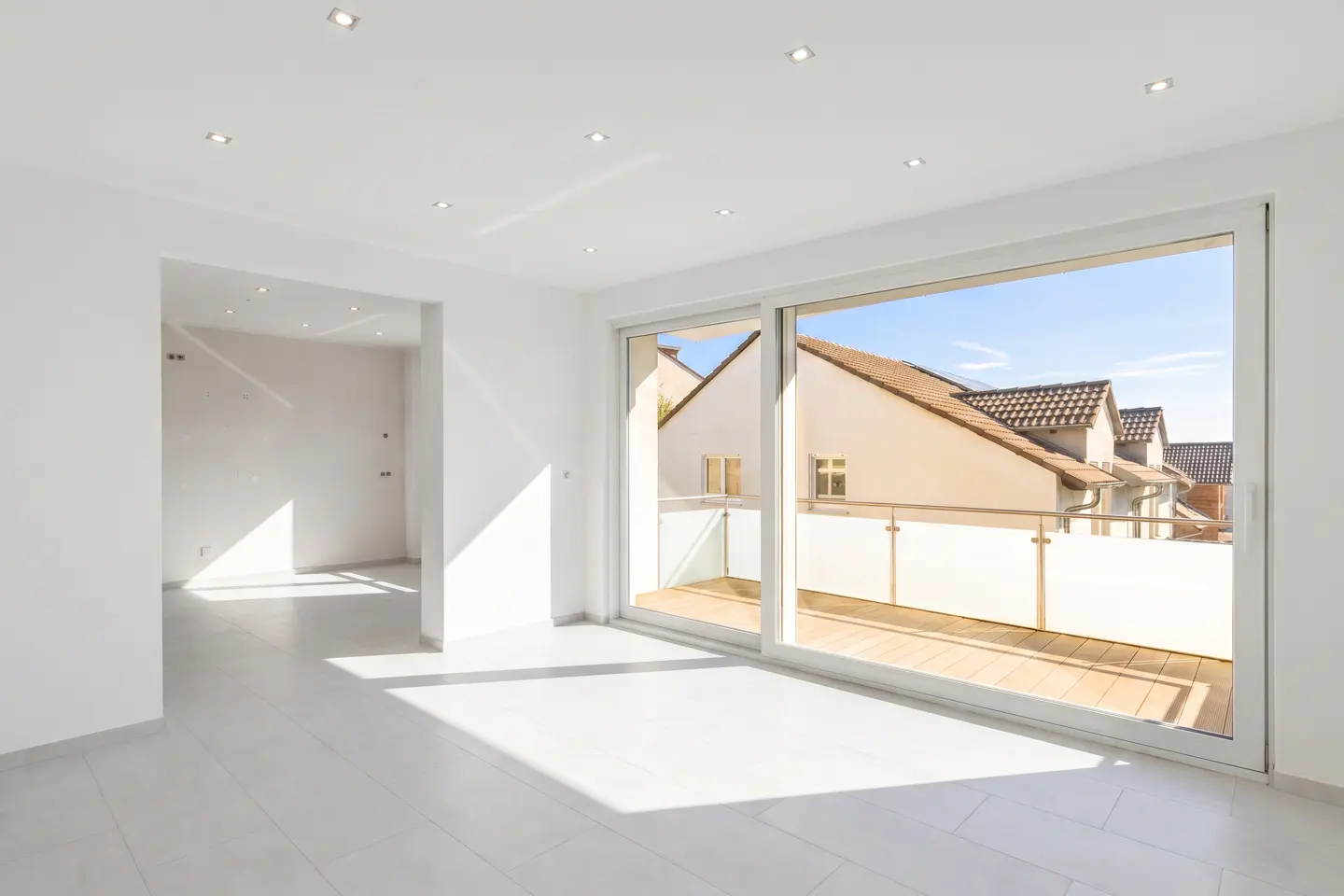 Bright, empty room with white walls, tile floor, and recessed lighting. Sliding glass doors open to a balcony with a view of houses.