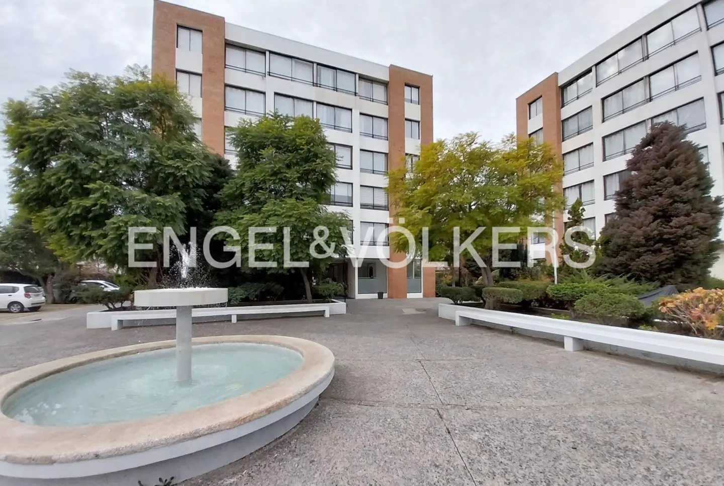 Exterior view of a white apartment building with a fountain in the foreground and the Engel & Volkers logo.