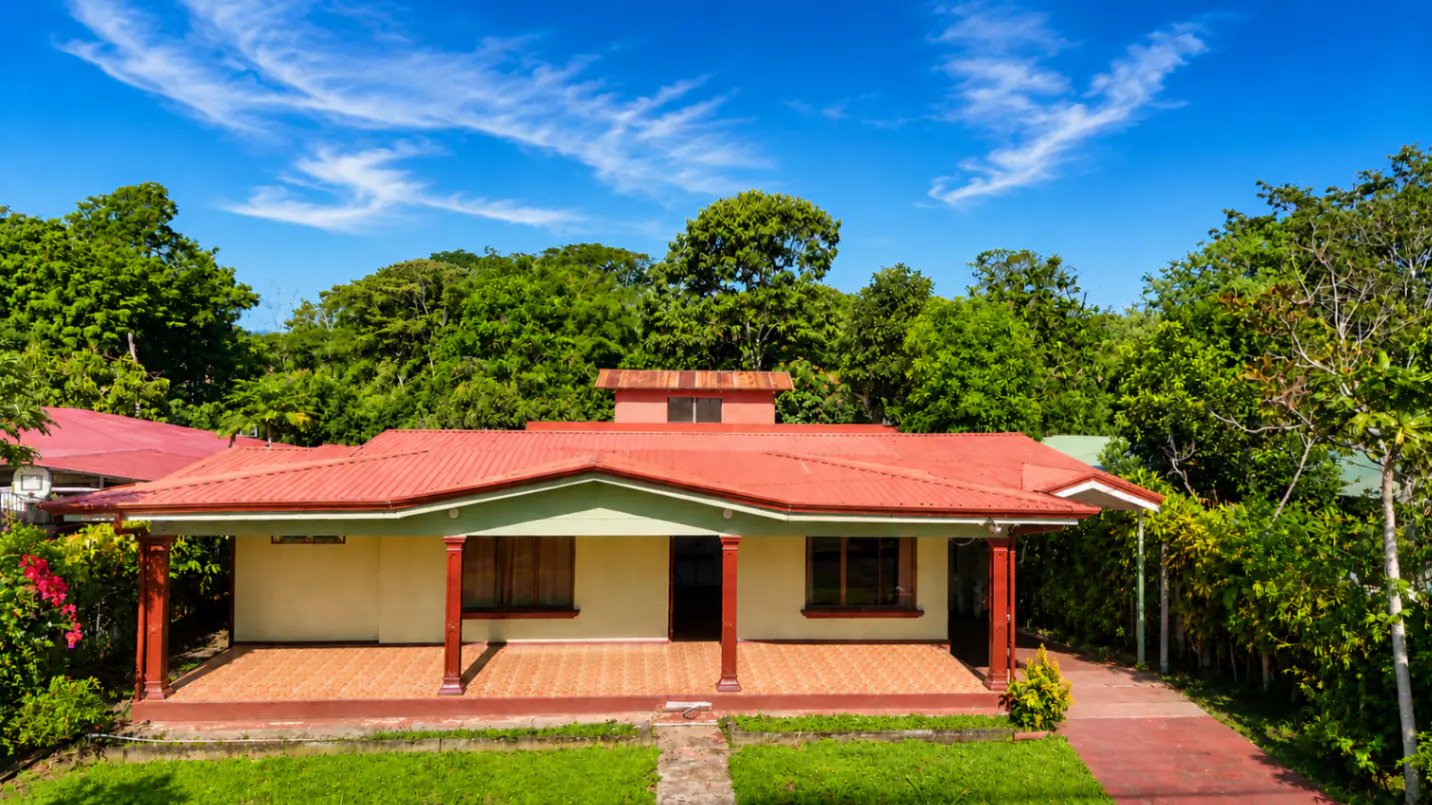 A tan house with a red roof sits in front of a lush green forest under a blue sky with wispy clouds.