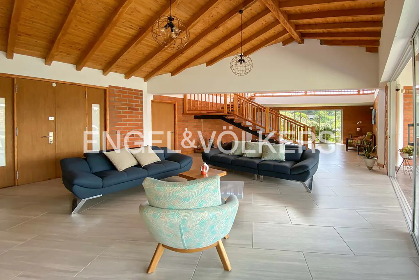 A bright living room with blue sofas, a patterned chair, and a wooden ceiling. A staircase leads to an upper level.