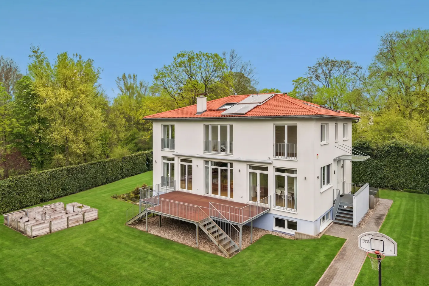 Two-story white house with a red tile roof, solar panels, and a wooden deck in a green lawn.