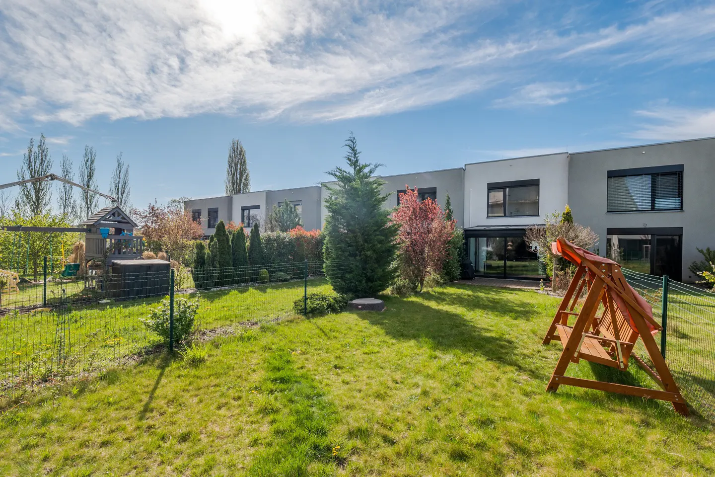 A backyard with green grass, a wooden swing set, and a playhouse. Modern gray townhouses are in the background.