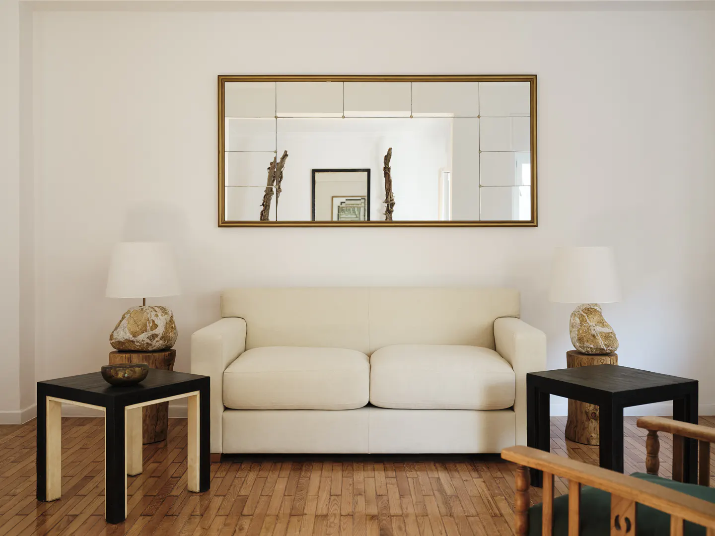 Living room with a cream sofa, black side tables, and lamps with stone bases. A gold-framed mirror hangs above the sofa. Wooden floors add warmth.