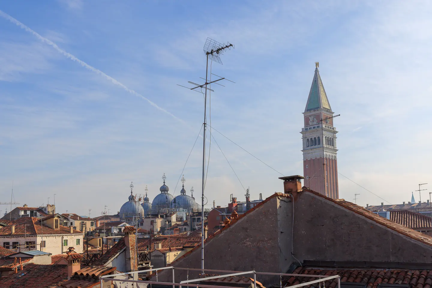 View of Venice rooftops with St. Mark's Basilica domes and Campanile tower under a blue sky with a contrail.