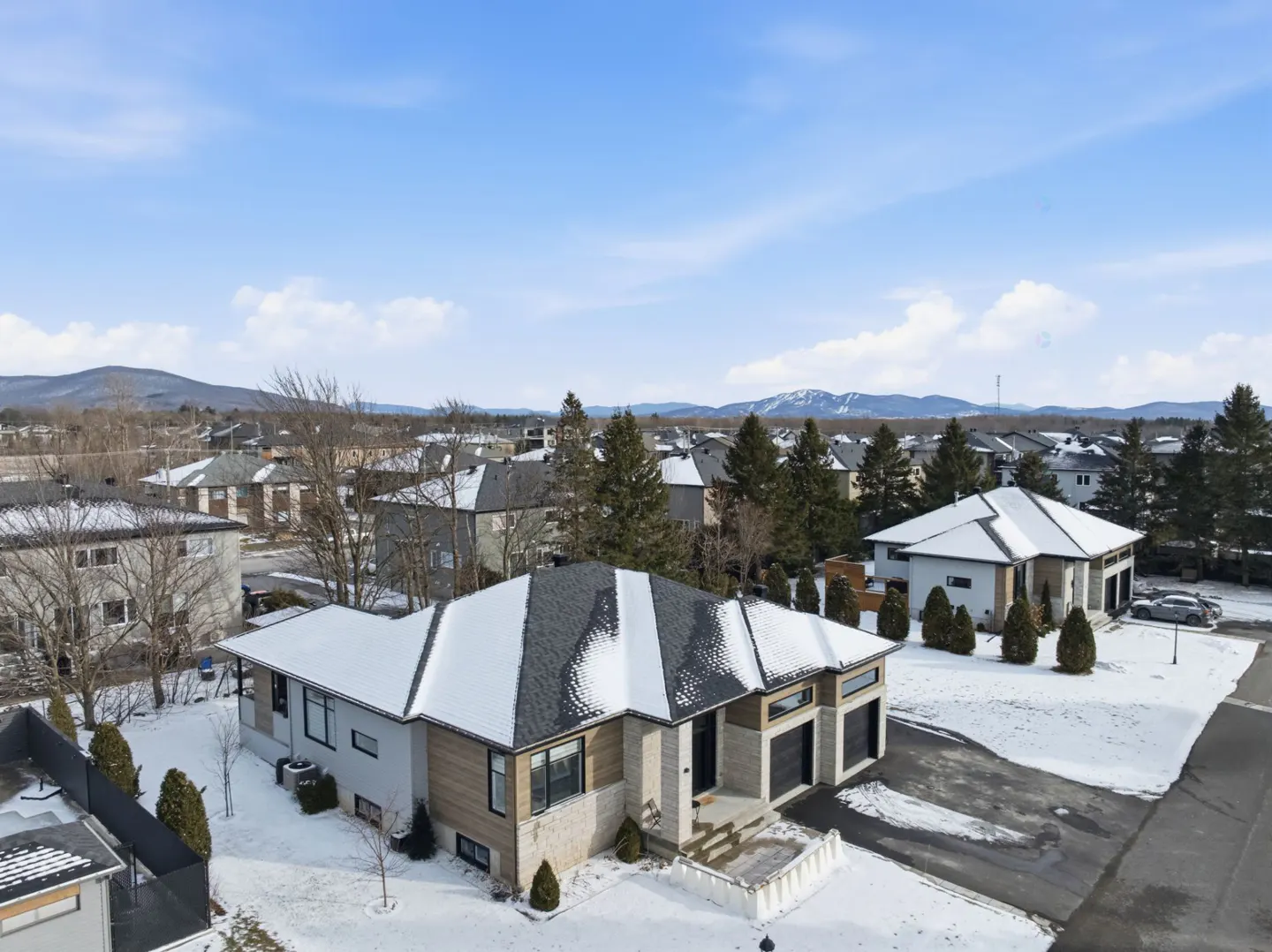 Aerial view of a modern, single-family home with a snow-covered roof in a suburban neighborhood during winter. Mountains in the background.