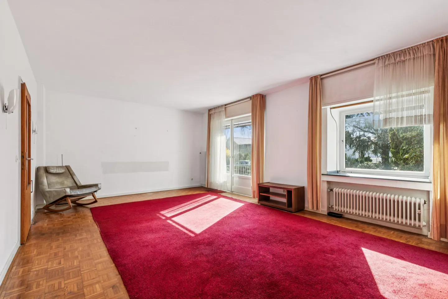 Bright living room with a large red carpet, a rocking chair, wooden floors, and windows with beige curtains.