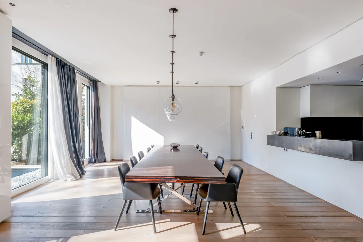 Bright, modern dining room with a long, dark wood table, black chairs, and a unique pendant light. Large windows let in natural light.