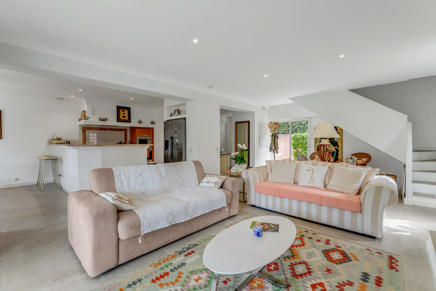 Bright living room with a beige sofa, a striped sofa with pink cushions, and a colorful rug. A white kitchen is visible in the background.