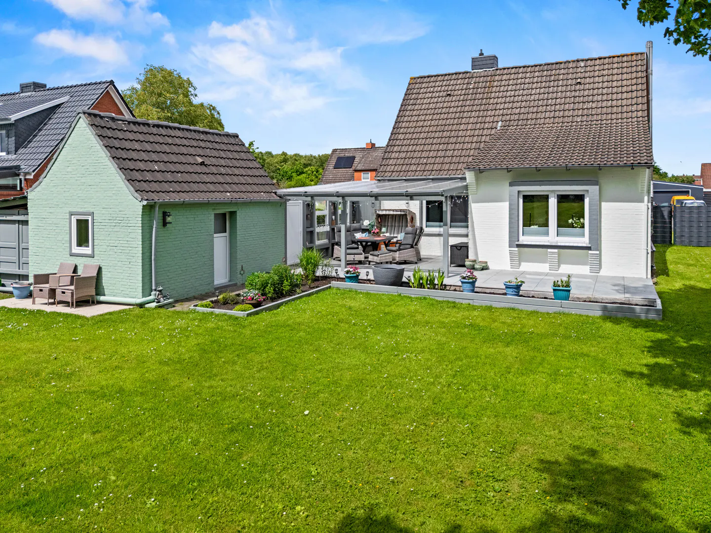 Backyard view of a white house with a patio, a green detached garage, and a large lawn under a blue sky.
