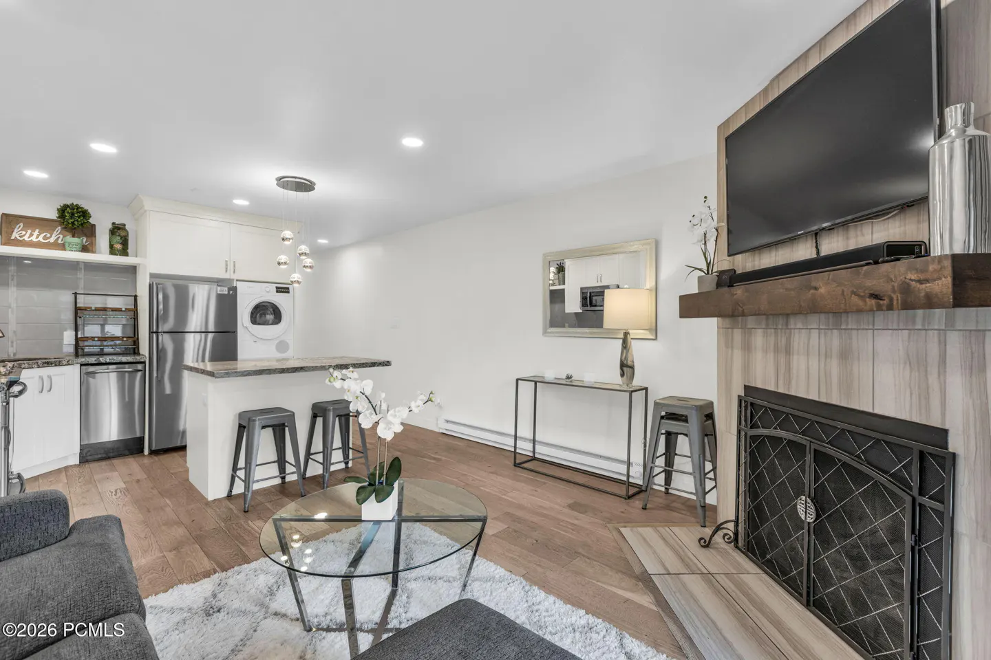 A modern living room with a kitchen, fireplace, TV, and gray sofa. The floor is wood and there is a white rug.