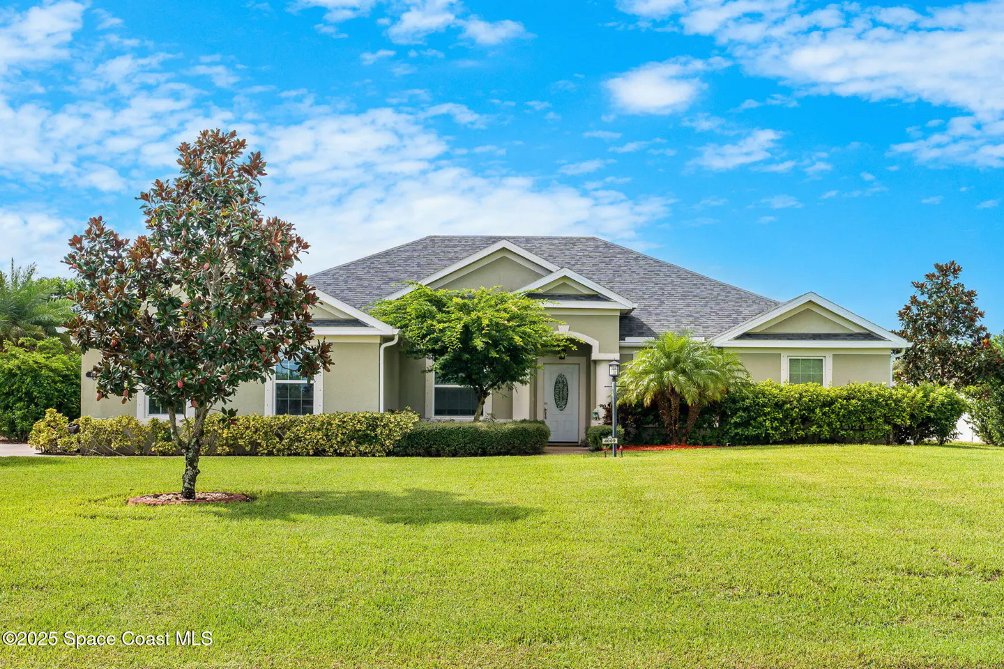 Single-story light green house with a gray roof, green lawn, trees, and blue sky with clouds.
