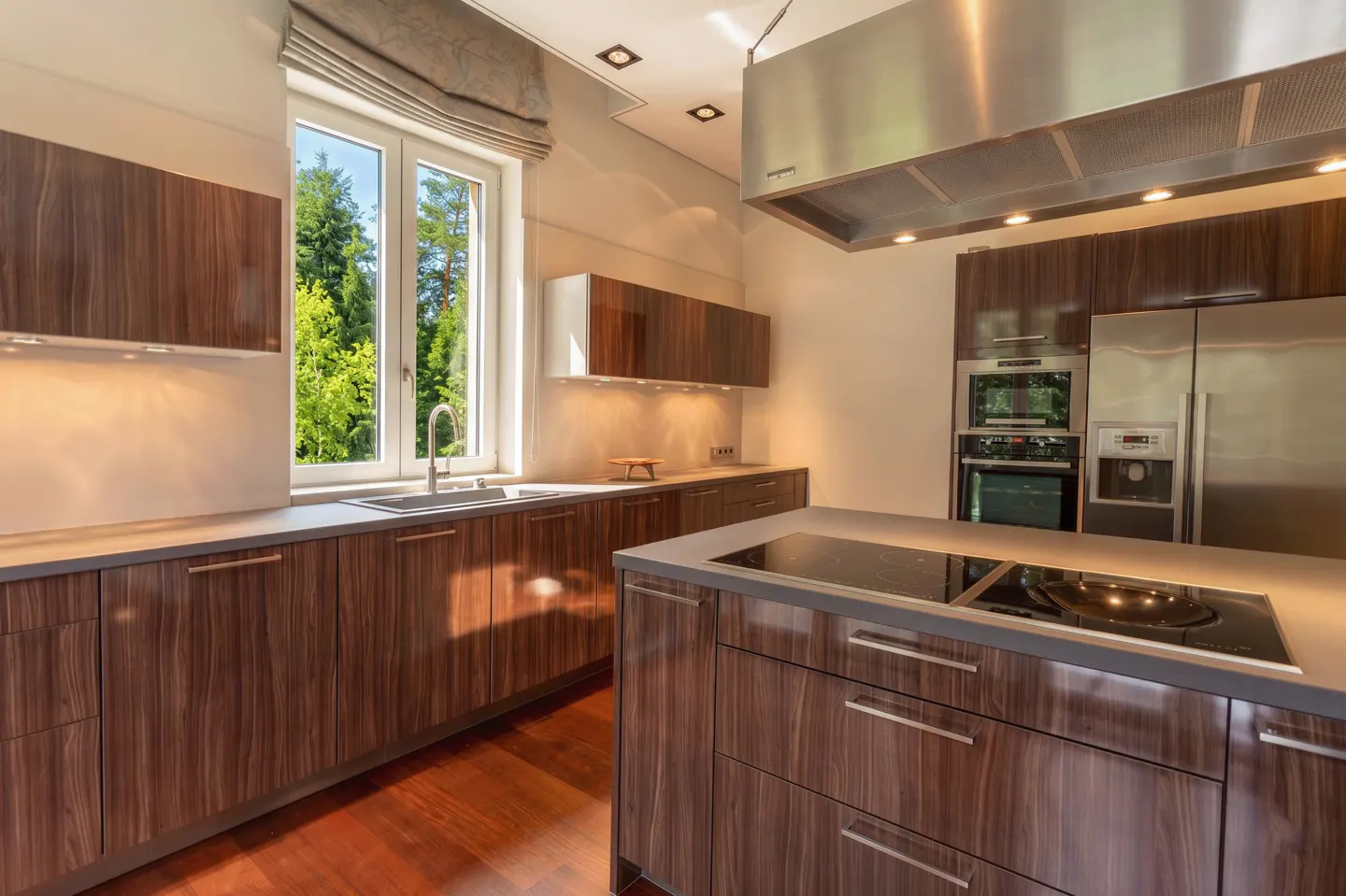 Modern kitchen with wood cabinets, stainless steel appliances, and a large window overlooking green trees.