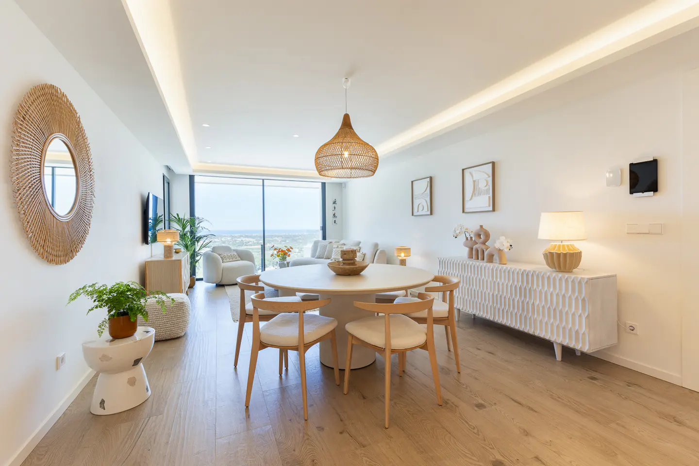 Bright, modern living room with a round table, six chairs, and a large window overlooking a city. Wicker mirror and pendant light add texture.