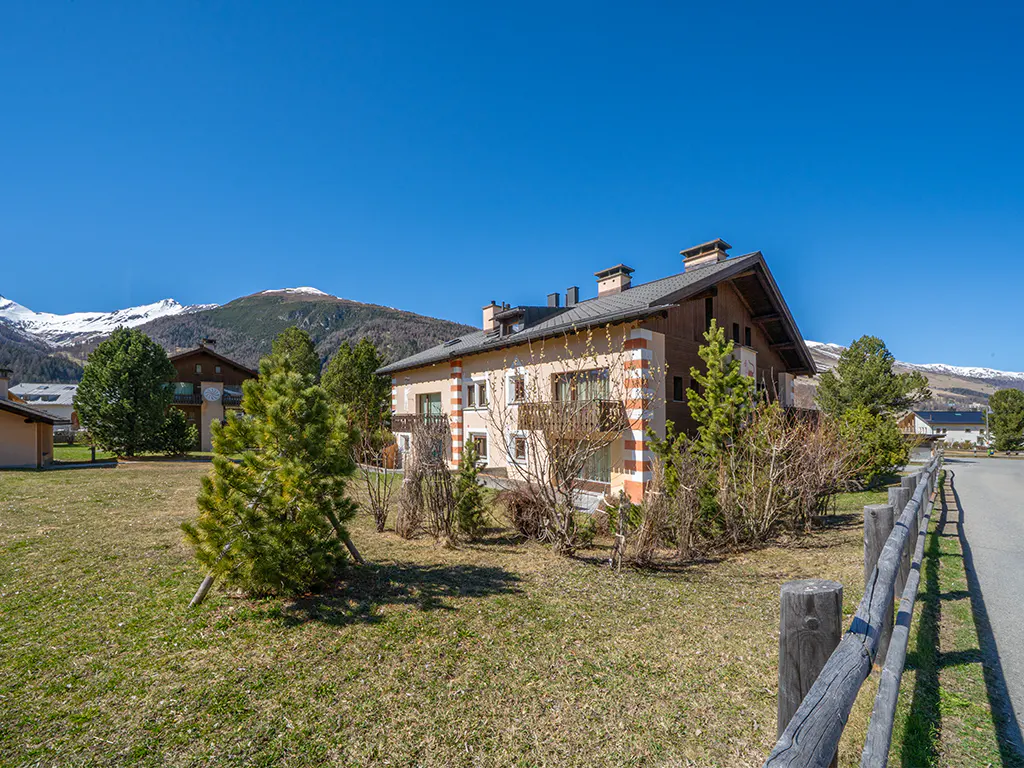 A multi-story chalet with tan and white striped corners sits in a grassy field against a backdrop of snow-capped mountains.