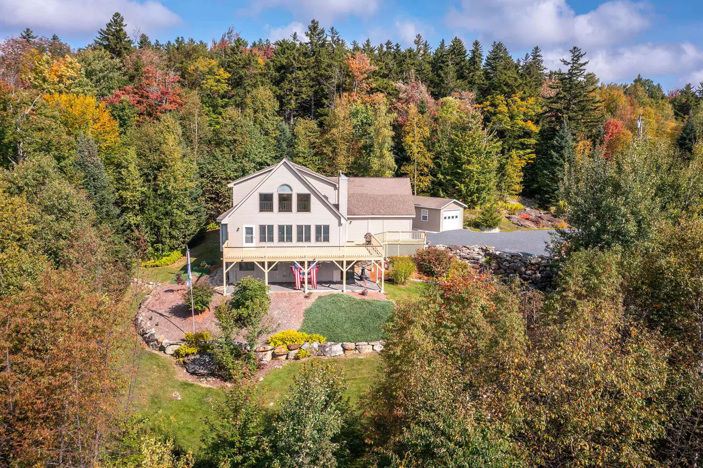 Tan house with a deck and American flags, surrounded by colorful fall foliage.